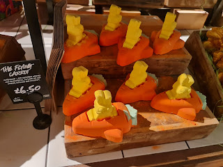 A group of orange rocket shaped bath bombs with a white small bunny shaped bath bomb in the middle on a light brown rectangular box next to black card that says the flying carrot in white font on a bright background