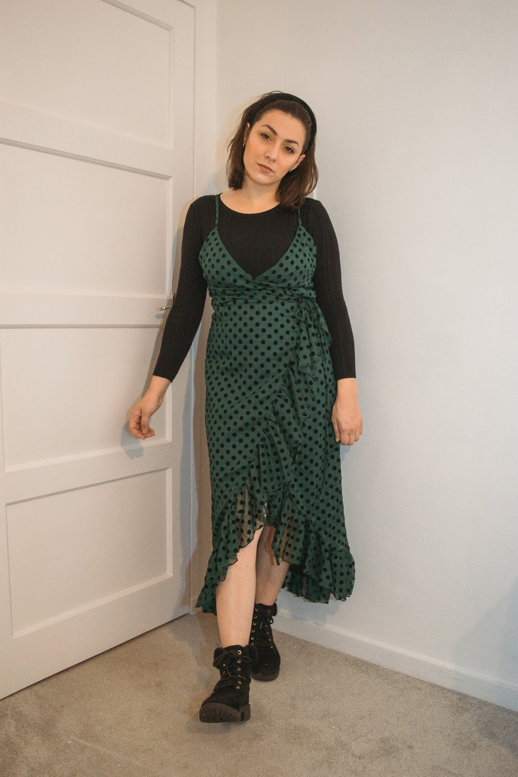 I am standing against a white background and I am wearing a black ribbed long sleeve top under a green frill polka dot dress that is a mid length. I am wearing black boots with fluffy trim.