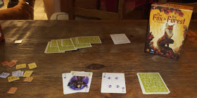 A game of The Fox in the Forest in progress. The box, with cover art depicting a fox sitting proudly atop a stone surrounded by flowering plants, stands near the draw pile, discard pile, and currently played cards. An array of cards, which are tricks taken by one of the players, sits nearby, along with a scoring summary and a pile of victory point tiles in 1, 3, and 6 point denominations.