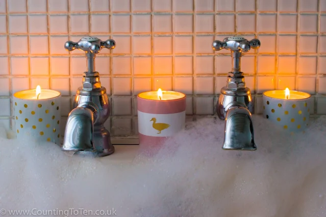 Two white candles with gold spots and a pink and white candle burning on the side of the bath with lots of bubbles