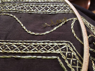 A close-up of gold-embroidered smooth brown silk in a wooden embroidery hoop, with two widely spaced borders of four lines of stitching each, with small diamonds dotted in the centers filling the space between the center lines of each border, and an undulating line of chain stitch through the wide space between borders. A short line of snipped threads, like tufts of seed fluff, spring from the line and point into the open curve described by it.