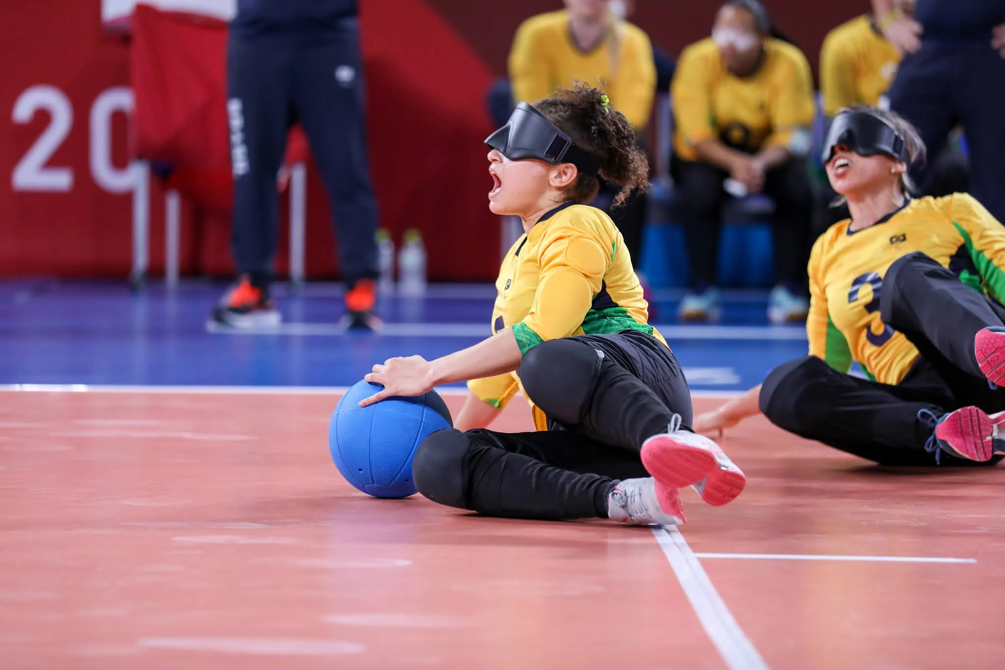 Jogadoras da seleção brasileira de goalball feminino, de uniforme amarelo, deitam no chão e arragam a bola