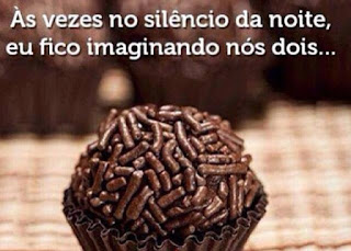 Foto em close de um brigadeiro em uma forminha de papel marrom, sobre uma toalha xadrez em tons de marrom. Ao fundo, imagem desfocada de vários brigadeiros. Acima do brigadeiro a frase em letras brancas: Às vezes no silêncio da noite, eu fico imaginando nós dois...