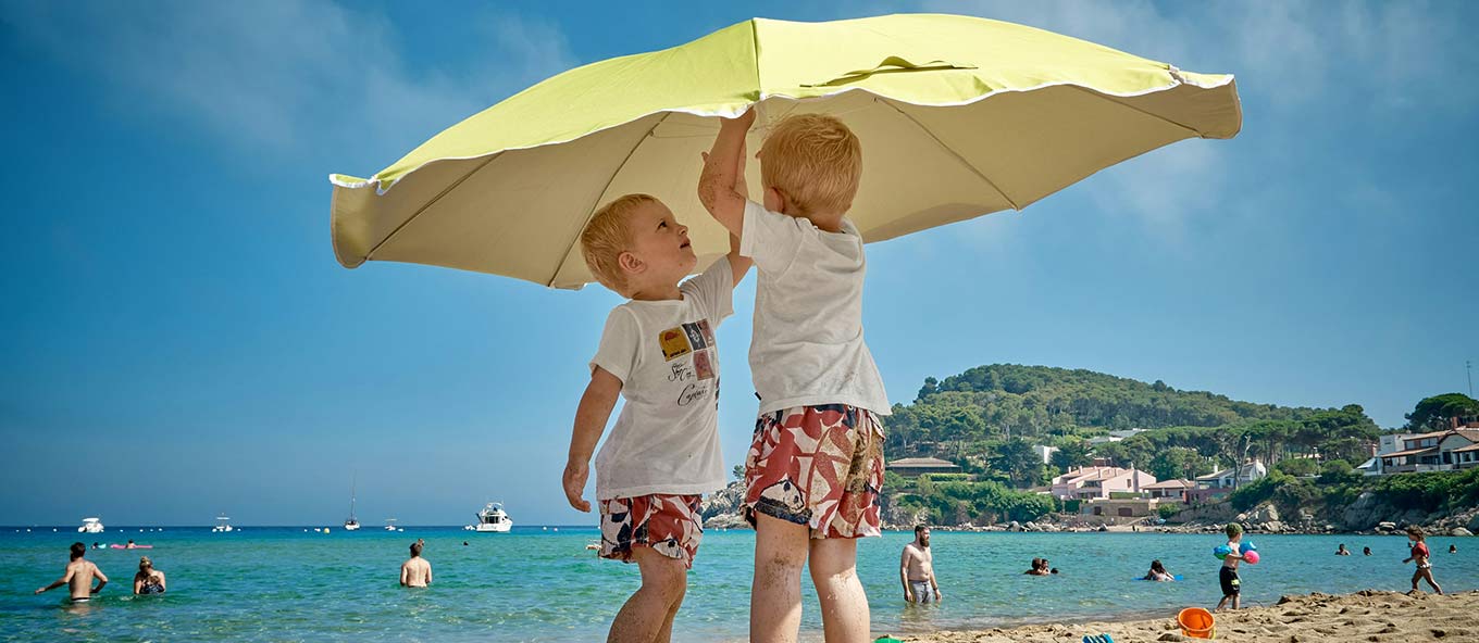 Two children in matching shorts and t-shirt putting up a yellow umbrella on a Spanish beach to protect themselves from the sun.