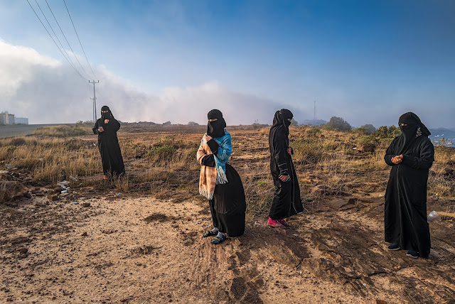 Four women in niqab and abayas stand separately in a rocky field
