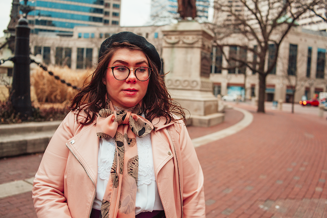 An outfit of a black beret, pink peplum faux leather jacket, vintage white pleated blouse, pink geometric skull print scarf tied in a bow, purple skater skirt, black tights and black boots.