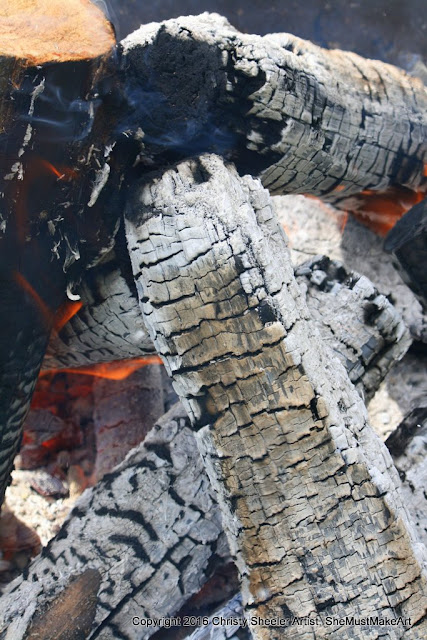 Another view of the campfire, black and white textures of wood logs turning to coals, with flames dancing around the edges of the wood.
