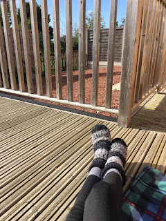 A persons legs and fluffy socks as the person lies on decking, with a patio and blue sky visible through the decking railings.