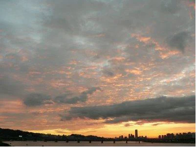A sunset view from Banpo Bridge facing Yeouido, Yongsan, and Mapo. The sky is streaked with reddish clouds, and the Hangang Bridge appears prominently in the foreground with the iconic 63 Building standing tall behind it. City silhouettes stretch faintly across the Han River.