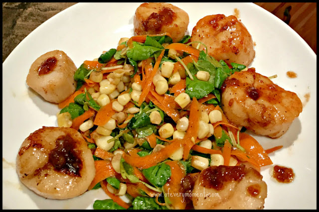 A colorful salad of corn, shredded carrots and greens surrounded by six caramelized scallops arranged on a white plate.