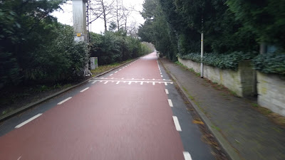 A red road with narrow black edge strips. There are trees left and right and a property wall to the right too.