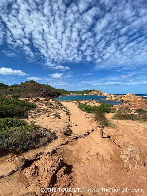 A red sandstone path leads to a golden sandy beach in a bay surrounded by sandstone cliffs and green hills, under a blue sky with a few white fluffy clouds.