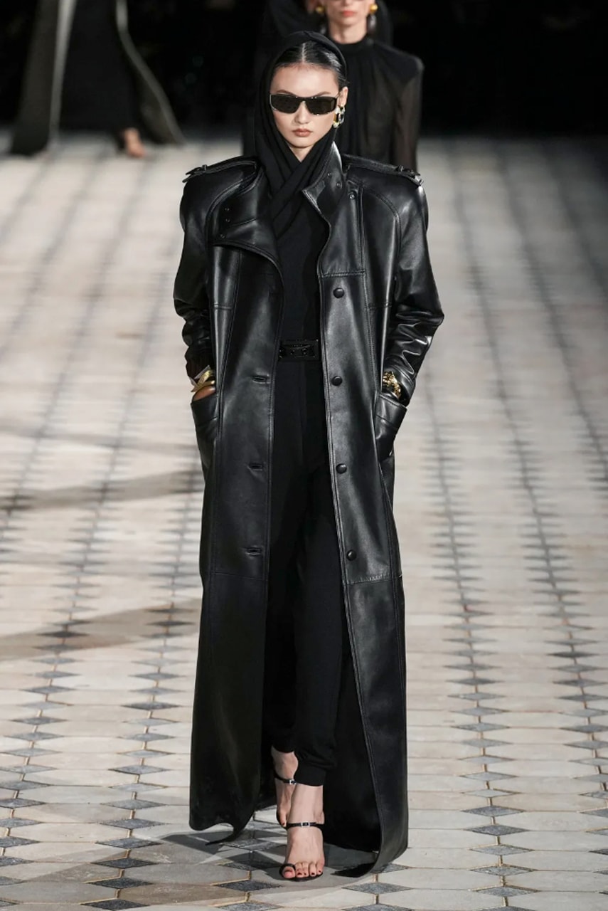 Woman with black hair in a bun and a middle part wearing black rectangular sunglasses, a black cotton scarf around her neck and over her head, silver oversized earrings, a black leather trench coat, a pair of black tapered skinny trousers, black strappy high heel sandals with dark bugundy nail polish on toes with her hands in her pockets walking on a black and white tiled floor