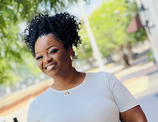 Kim Robinson is smiling at the camera. She is outside and it is a bight and sunny day. You can see trees in the background. She is wearing a white shirt.