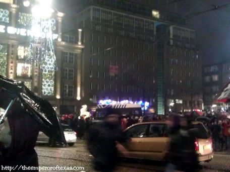 Dam Square. Riot police guard the place at all entrances and stand firm in the middle of Dam Square. 11 minutes to THE NEW YEAR 2000 and 13(LIVE caption delivered to Fb: “Police guard DAM SQUARE as more and more serious explosives are being heard. some very loud explosions, shiver through me.11 minutes to NEW YEAR 2013.”)