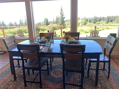 Dining table, modern, with six chairs. Flowers on the table, persian rug beneath. Windows behind chairs show view of Alaska landscape, with pines, at mid-day.