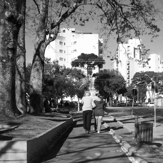 Fotografia P e B. No centro da imagem uma pista de caminhada numa praça ensolarada. De costas, caminham na pista, de braços dados, uma mulher idosa de cabelos curtos e menina de cabelos longos e presos. Usam agasalhos, moletons, tênis. À esquerda da pista, canteiro alto com gramado, três troncos grossos de árvores enfileirados e à direita, canteiro baixo com gramado, lixeira, calçada e via pública. Ao fundo, árvores, copas arredondadas, uma araucária, veículos estacionados, prédios altos, céu sem nuvens.