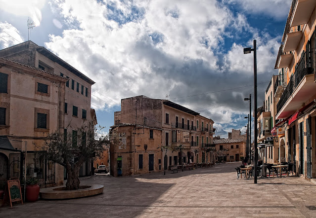 Das foto zeigt den Hauptplat, also den Placa Mayor von Santantyi auf Mallorca. Es zeigt urbane und typische. mallorwuinische Bauten in Sandstein. Die Straße ist gepflastert und der Ort strahlt eine große Gemütlichkeit aus.