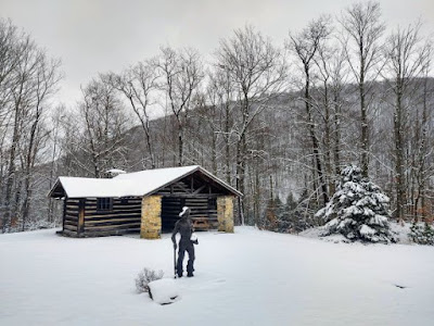 Snowy landscape at PA Lumber Museum with stone and log cabin and statue of a CCC worker. The statue is black and the worker is shirtless, wearing a brimmed hat and hold an ax with its head resting on the ground.