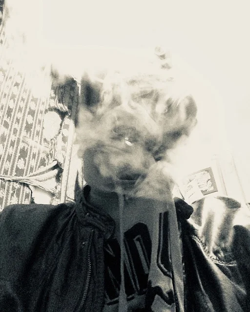 Black and white photograph from inferior POV Oregonleatherballs standing superior wearing blue fashion leather jacket blowing thick smoke that covers face