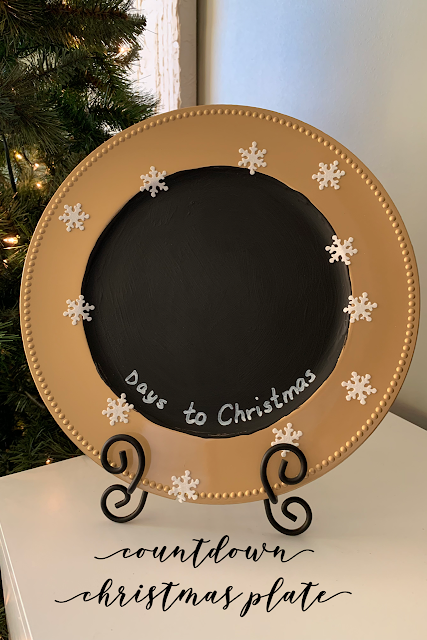 Round gold plate charger with black chalkboard paint in the center, with the words Days to Christmas written in white sharpie paint on the bottom and decorated with white snowflakes.