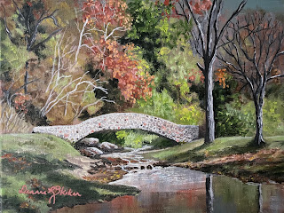 An acrylic painting by Deanna Skokan of a stone bridge across a small creek. The grass on either bank is green, and the foliage of the trees around is turning orange from the fall. There are two bare trees on the right side.