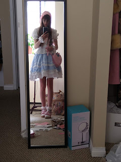 Lolita girl wearing a blue skirt, white blouse and pink accessories standing in a messy room in front of her mirror.