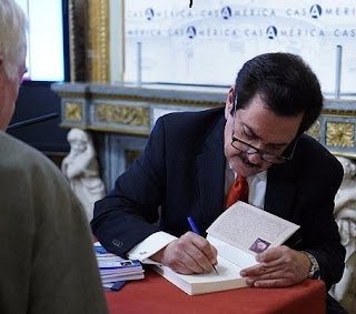 En la foto, el escritor Raful está sentado en una mesa con mantel rojo, firmando un ejemplar de su libro. Tez clara, pelo y bigote negros. Usa lentes. Viste traje negro, camisa blanca y corbata roja. Un hombre observa.