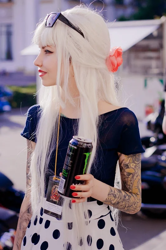 Chica de pelo largo blanco, esta paeando con una bebida, lleva tatuajes en el brazo izquierdo