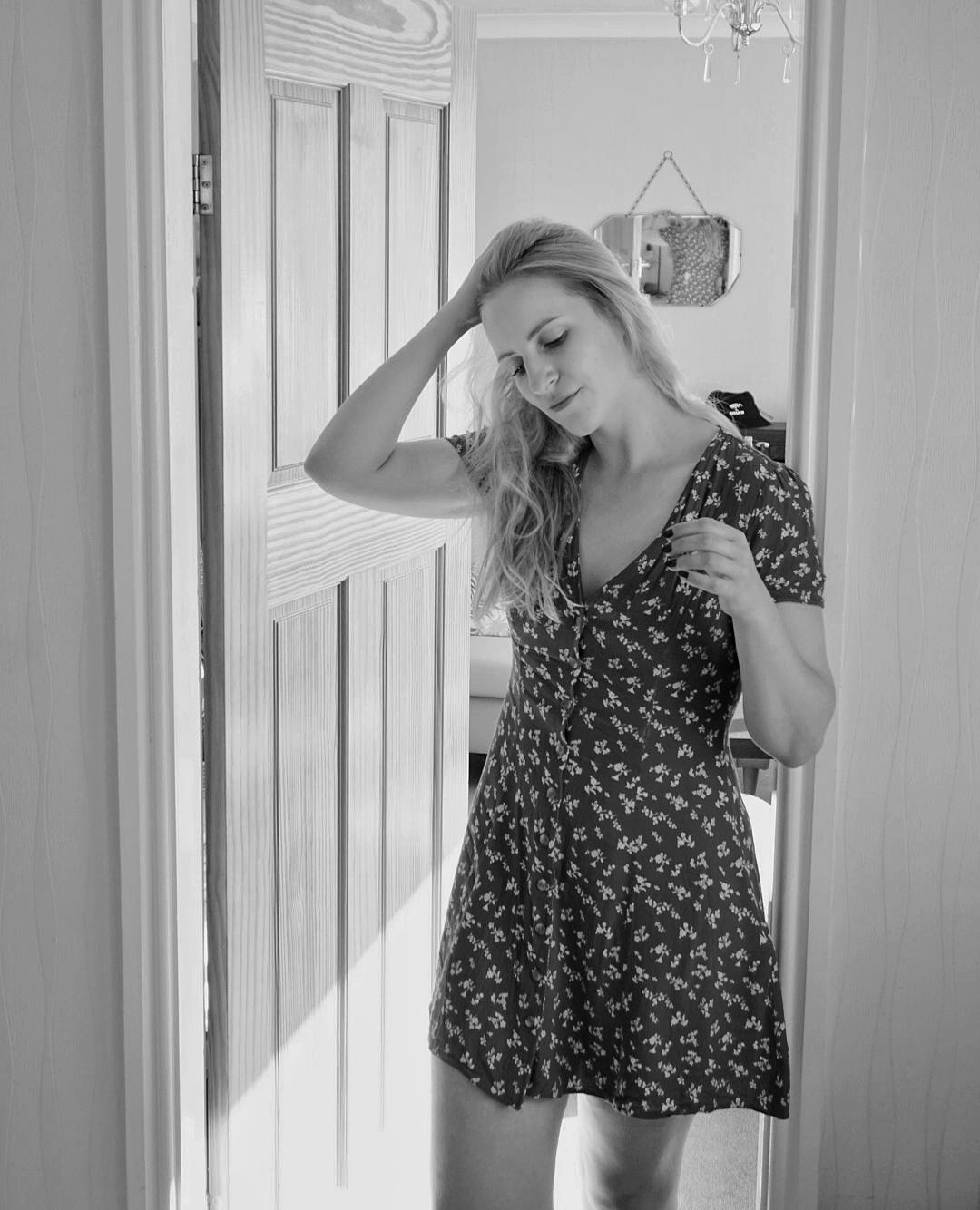 black and white photograph of a woman in a short floral dress standing in a doorway, running her hand through her hair