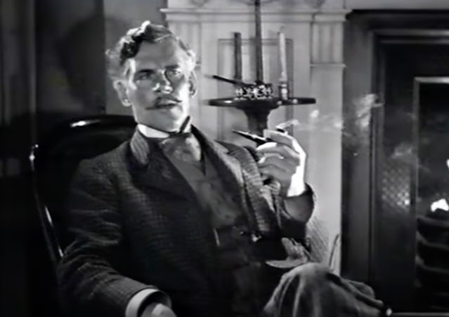 Walter Huston sitting in a chair by a fireplace with a lit pipe in his hand, dressed for his role in the 1939 movie The Light That Failed set in the late 1800s.