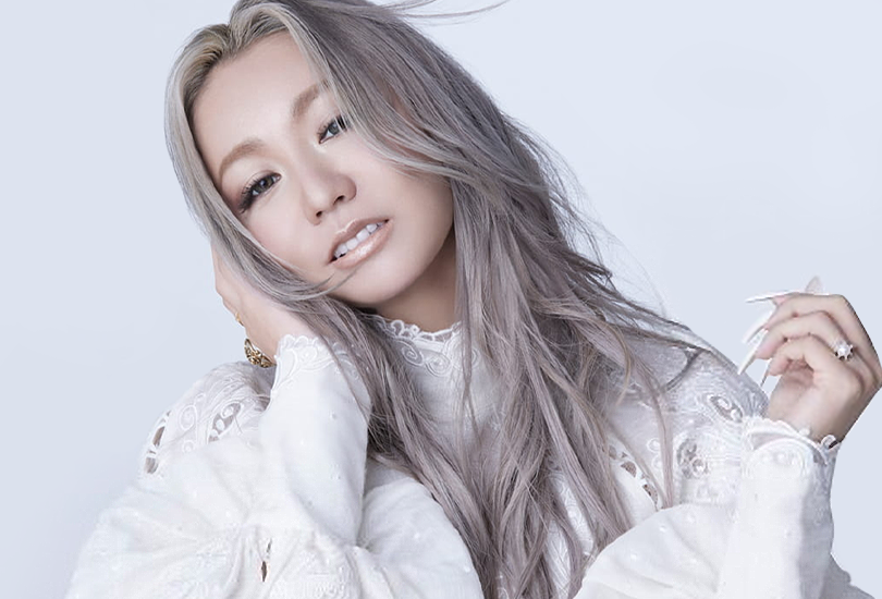 A shot of Kumi Koda, posing with her hand clasping her face, whilst wearing a white dress with lace detailing, with her hair down and coloured shades of grey.