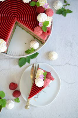 Himbeer-Dirndlmousse-Torte mit Brennnessel-Biskuit von oben fotografiert – zu sehen ist die tiefrote Himbeerspirale und rundherum Schlagoberstupfen, frische Himbeeren und Himbeeren aus Dirndlmousse, Baiser, Blätter und Blüten. Ein Stück der Torte ist herausgeschnitten und auf einem kleinen Teller platziert.