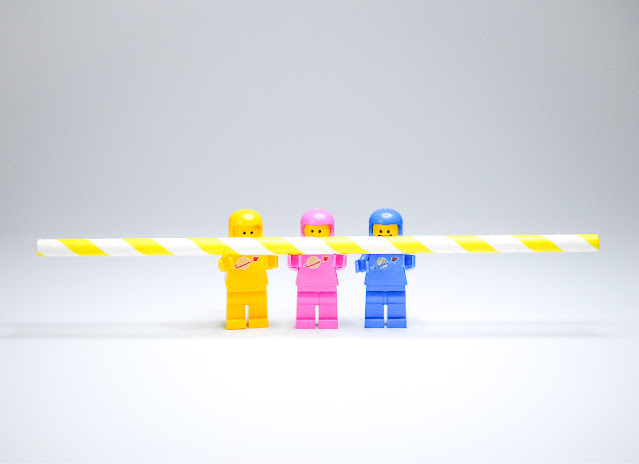 Three Lego mini-figs of different colours are working together to hold a straw.