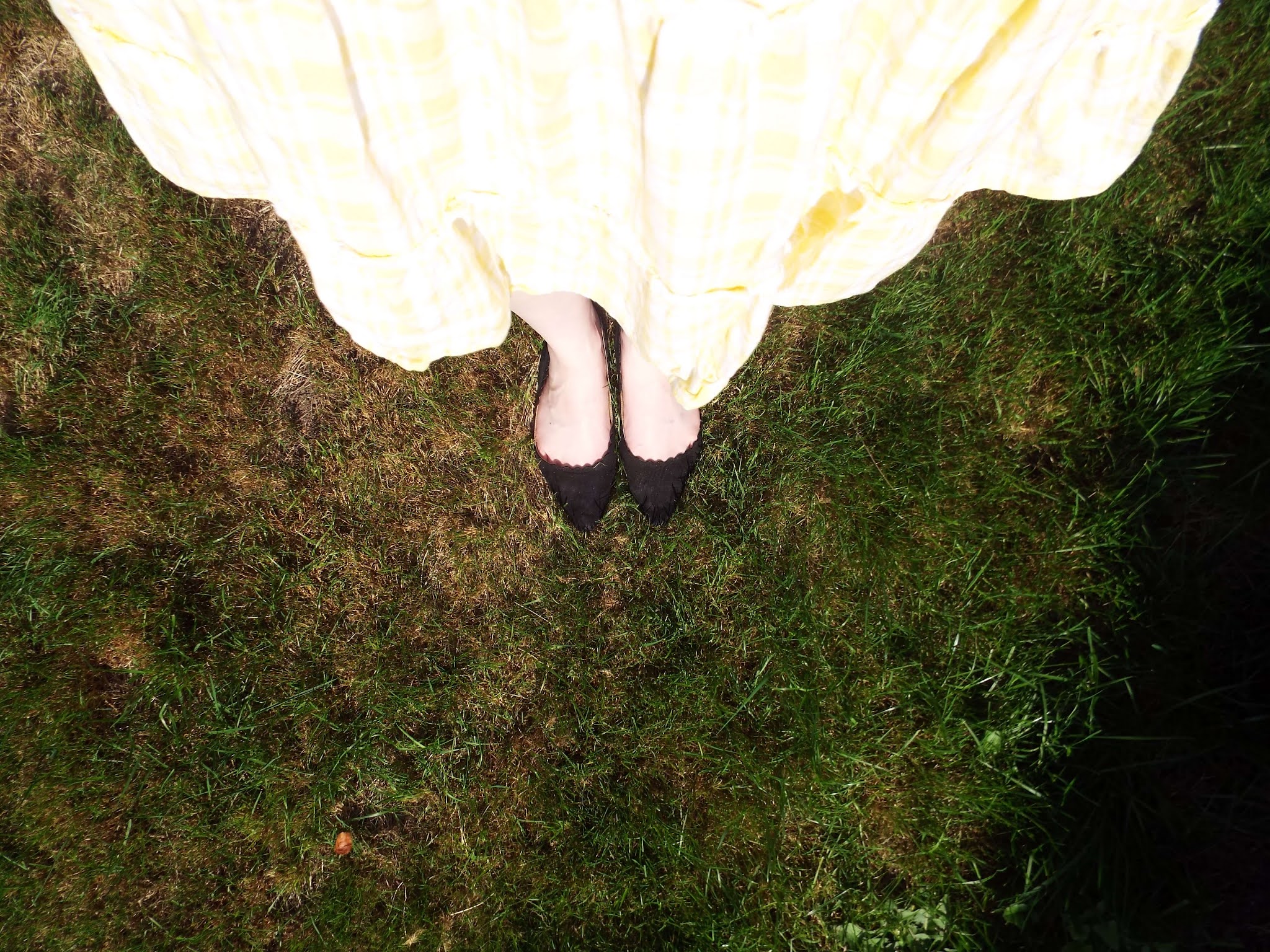 Picture looking down to my feet on the grass, whilst stood up, showing my pointed toe, scalloped edged black slip-on shoes and yellow gingham Summer dress, floating in the breeze.