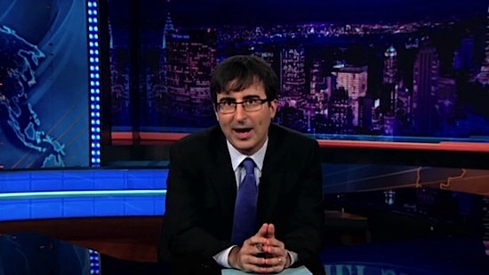 John Oliver, a white man in glasses wearing a dark suit and blue tie with tousled dark hair, on set