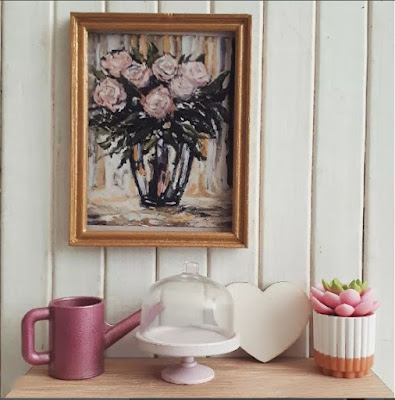 One-twelfth scale modern miniature shelfie containing a framed print of flowers, a watering can, cake stand and cover, wooden heart and potted succelent.