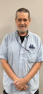 Man with dark hair pulled back and a gray beard wearing a light blue short-sleeved collared shirt with "U.S. Brig Niagara Crew" emblem