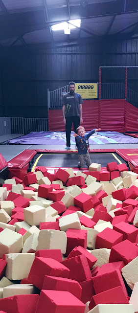 Little boy bouncing from a trampoline into a foam pit as he Daddy watches in the background