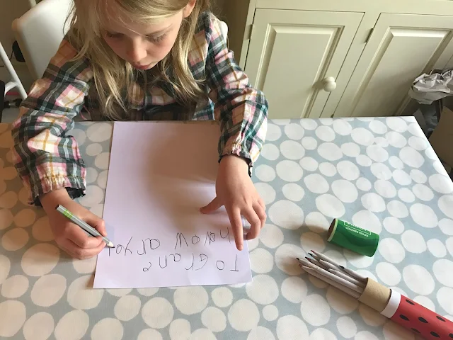 M is still writing the letter with the write size pencil. The Froot colouring pencil box is open next to her. She has written "To Grane howow ar you"