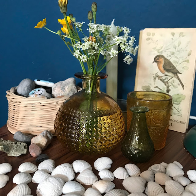 Zwei Glasvasen in grün und gelb auf einem braunen Beistelltisch vor einer dunkelblauen Wand. In der größeren Vase sind Wiesenblumen, davor und daneben liegen Steine und Muscheln. Hinter den Vasen ein oranges Glas-Kerzenlicht und ein altes Buch mit einem Vogel auf dem Cover.