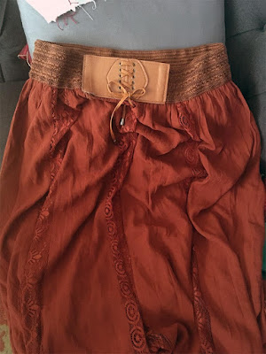 A rust-red skirt with narrow vertical panels of lace, a dull-orange elastic waistband, and a faux leather panel with corset-style lacing at the front center of the waistband.