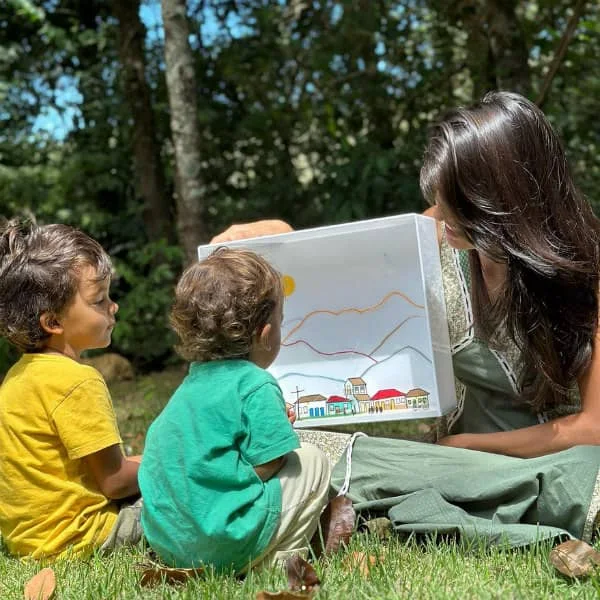 two young children look at miniature paper cutting diorama held by smiling young woman