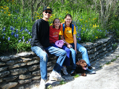 My family sits on a low stone wall in front of a bank of blue and yellow wildflowers; Alfie is lying down at Charissa's feet