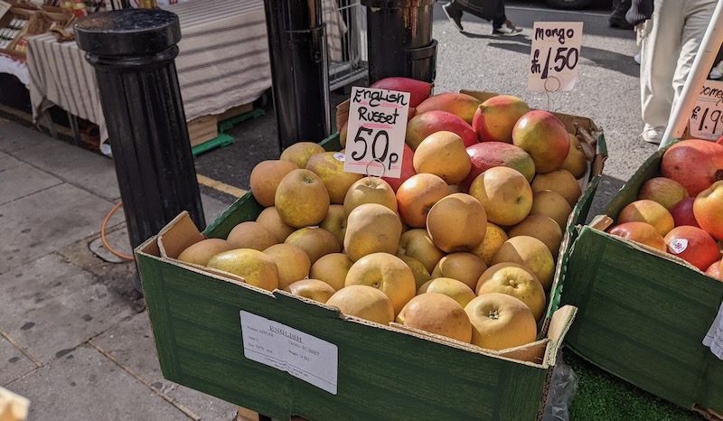 A coarboard box full of ruseeted apples, with a hand0letered sign that says "English Russet 50p." The box also has a printed label that says "English Russet."