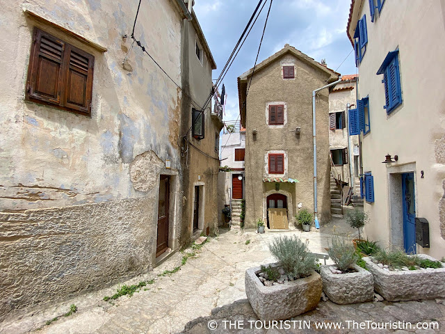 Three-storeyed houses decorated with flower pots and blue shutters on a narrow cobble-stoned lane.