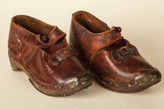 A child's pair of red-brown leather clogs with a nailed wooden sole. They have an ankle bar with a small brownb button, and a small decorative steel buckle. There is brass on the toe tip. They have horseshoe irons on the sole and heel.