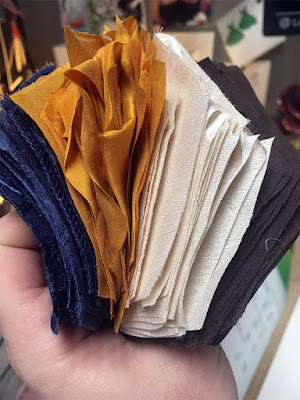 A white hand holding a four-colored stack of large silk hexagons, seen on edge. From left, they're medium blue, gold, cream, and dark brown.