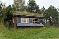 A small wooden house with a green roof and solar panels, located in the countryside. There is a porch in front of the house and a garden on one side.
