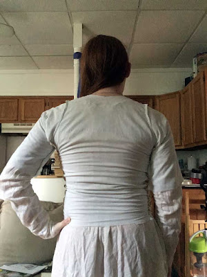 A young, redheaded white woman from behind, in a small room with wooden kitchen cabinets in the background. She's wearing a white chemise with long, tight sleeves and a palest aqua bodice with half sleeves, and has her left hand propped on her hip. The lower back is heavily wrinkled horizontally, and a small bubble of fabric puckers off her left shoulder where the pose distorts the bodice.
